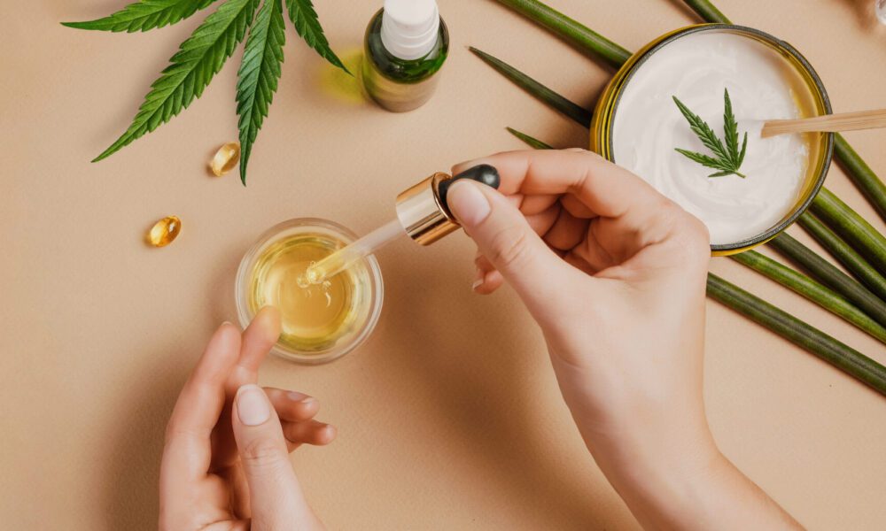 Pipette with CBD cosmetic oil in female hands on a table background with cosmetics, cream with cannabis and hemp leaves, marijuana. Flat lay, top view.
